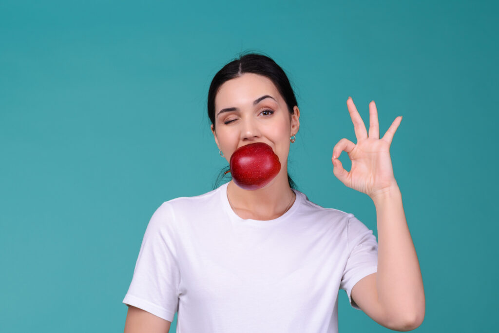 Young attractive woman holding a red apple in her mouth and shows ok sign. Healthy vegetarian food concept.